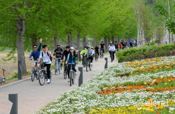 자전거를 탄 사람들이 자전거 도로를 달리며 손을 흔들고 있다. 자전거길 옆에는 꽃들이 화사하게 피어 있다. / 증평군 제공